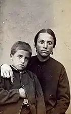 Portrait en photographie sépia d'une femme habillée simplement, au visage rehaussé de deux boucles d'oreille, et posant son bras droit sur l'épaule de son fils, au regard hagard.