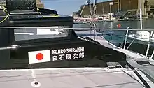 Côté bâbord de la « casquette » noire du bateau, sur laquelle figure le drapeau japonais blanc à gros point rouge, à droite duquel on peut lire en blanc « Kojiro Shiraishi » et, en dessous, ce même nom en caractères japonais.