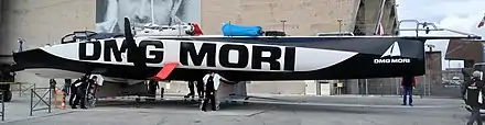 De profil, bateau blanc et noir, non mâté, mais avec ses foils, tiré sur un chariot. Des membres du Team sécurisent l'opération.