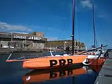 Voilier orangé et blanc vu de trois quarts avant, amarré au ponton.