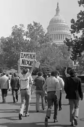 Un manifestant tient au-dessus de sa tête une pancarte où est inscrit « Impeach Nixon ». D'autres personnes marchent comme lui en direction du Capitole.