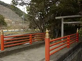 Photo couleur d'un pont en ciment (premier plan), surmonté de balustrades faites de bois peint en rouge vermillon. Un portique (à droite), marque l'entrée d'un lieu de culte shintō.