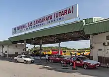 Photographie d'un terminal de bus dont le nom est « Terminal Bas Bandaraya (Utara) ». Deux taxis et une voiture civile peuvent être vus au premier plan et quatre bus jaune et rouge à l'arrière-plan.
