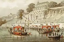 Lithographie en couleurs représentant une église en haut d'une falaise dans laquelle est située une grotte et, en contrebas, des embarcations décorées festivement, traversant un cours d'eau très large.