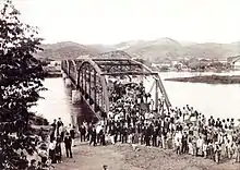 Une vieille photo montrant une foule de gens au premier plan avec un pont en fer qui enjambe une rivière en arrière-plan