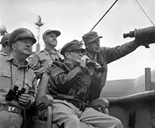 MacArthur est assis et tient une paire de jumelles. Quatre autres hommes en uniforme et portant également des jumelles sont derrière autour de lui.