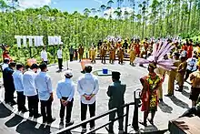 Rassemblement circulaire de plusieurs dizaines de personnes autour d'un pavage au milieu d'une végétation tropicale. En fond, les mots TITIK NOL.