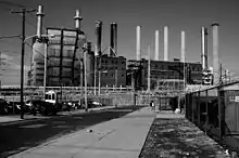 Photo en noir et blanc d'un paysage industriel. On trouve au premier plan une route qui conduit à des usines entourées de clôtures. Il y a une personne au milieu de la route.