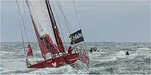 Voilier rouge et blanc, vu de l'arrière, sur l'océan. Le skipper se tient à l'arrière.