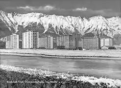 Photo en noir et blanc. À l'avant, on voit la rivière, puis le village olympique, un ensemble de grands bâtiments, et enfin en fond les montagnes enneigées.