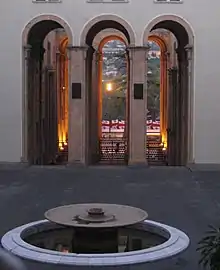 Fontaine circulaire, éteinte, au premier plan. Au deuxième plan, trois ouvertures hautes et arquées. À travers, on aperçoit au loin des drapeaux géorgiens blancs et rouges.