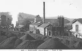 Photo noir et blanc montrant un complexe industriel avec bâtiments, trémies, cuves et cheminées entourés de terril, l'air est envahi de vapeur et fumées.