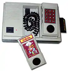 console quadrangulaire grise avec deux manettes en forme de combiné de téléphone. Une cartouche du jeu BurgerTime est posée dessus