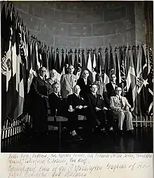  photo en noir et blanc montrant les dix membres du comté scientifique du congrès sur deux rangs, devant un rang assis de quatre hommes et au second plan un rang debout de six hommes, au fond en demi cercle les drapeaux des nations participantes au congrès