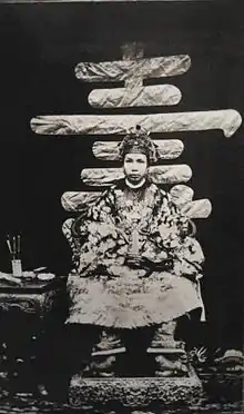 Portrait photographique d'un très jeune homme asiatique, en habit traditionnel richement décoré, assis sur un trône.