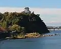 Photo couleur montrant un château au sommet d'une colline boisée surplombant un fleuve.