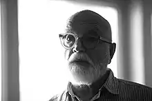 Portrait en noir et blanc d'un homme à la barbe blanche et aux lunettes rondes, dont la partie gauche du visage est dans l'ombre et la partie droite dans la lumière.