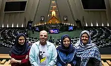 Photographie en couleur représentant un homme et trois femmes, ayant tous le visage vitriolé. À l'arrière plan on peut voir une grande salle d'assemblée.