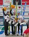 trois jeunes femme souriantes en bonnet aux couleurs de leur sponsor et vêtements de sport d'hiver sur un podium, tenant verticalement leur ski de saut dune main, et un bouquet orange de l'autre