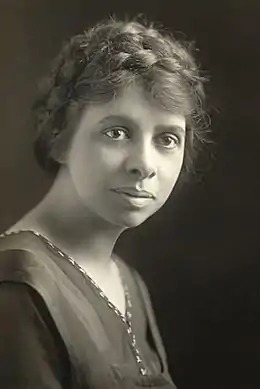 Portrait de jeune femme, regard caméra, volontaire. Cheveux rassemblés en nattes sur la tête, vêtements simples.