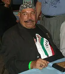 Homme corpulent aux moustaches en pointes assis à une table. Il porte un T-shirt noir, une veste noire et un bandana à effigie du drapeau américain.