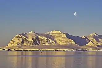 Photographie en couleurs d'un paysage montagneux faiblement éclairé, avec la lune visible en haut et à droite.