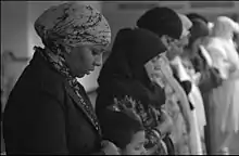 Photographie en noir et blanc d'une rangée de femmes en prière, les yeux fermés, la tête baissée et les bras croisés.