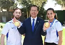 Un homme en costume se tient au milieu et montre les médailles d'or d'un homme et d'une femme habillés en tenue de sportifs.
