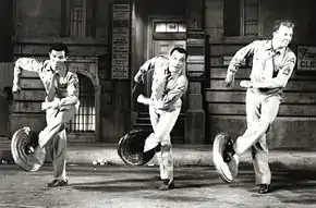 Trois hommes avec des couvercles de poubelles sur un pied chacun, en plein numéro de danse.
