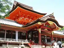 Photo couleur de la porte d'entrée à deux étages de l'enceinte du bâtiment principal d'un sanctuaire shintō, structure en bois laqué de couleur vermillon dont la toiture est en chaume. Le mur d'enceinte, de part et d'autre de la porte, forme un corridor dont la toiture est faite de tuiles grises. L'arrière-plan est constitué d'un ciel bleu lumineux.
