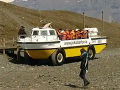 "Véhicule jaune et blanc a l’arrêt au sec devant un ponton, avec une vingtaine de personnes à bord portant des gilets de sécurité, dans un paysage minéral"