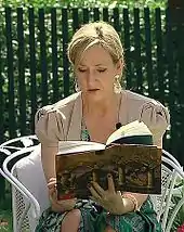 Une femme blonde aux cheveux attachés est assise en extérieur sur une chaise de jardin blanche. Elle est légèrement penchée en avant pour lire le livre qu'elle tient dans les mains