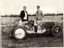 Photo de deux hommes se tenant de pied derrière une voiture à la carrosserie chiffonée.