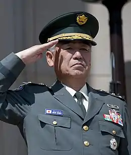 Le général Ryoichi vu de face faisant un salut militaire. Il porte un uniforme et une casquette de parade. Il arbore des décorations militaires à gauche.