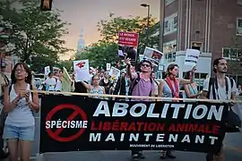 Nombreux manifestants antispécistes dans une rue de Montéral, avec plusieurs pancartes. L'une clame "Le monde est vegan ! Si vous le voulez." Sur une banderole portée à l'avant de la marche il est écrit "Spécisme : Abolition. Libération animale maintenant. Mouvement de libération."