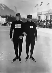 Photographie en noir et blanc de deux patineurs à l'arrêt au centre de la glace, se tenant pars le bras, portant une tenue sombre et un bonnet ainsi que le drapeau finlandais sur la poitrine.