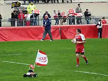 Un joueur de rugby entre sur le terrain en portant un ballon, précédé par une voiture télécommandée équipée d'une peluche de taureau et d'un drapeau.