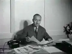 Photographie en noir et blanc d'un homme assis à son bureau tenant un journal dans ses mains.