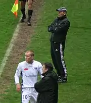 Trois hommes debout au bord d'un terrain de football, dont un footballeur et deux personnes en survêtement.