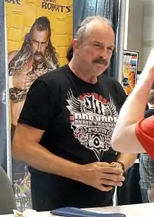 homme de 58 ans moustachu aux cheveux gris et front dégarni portant un Tshirt noir debout devant une affiche de catch