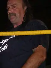 Un homme brun et moustachu portant un t-shirt noir.