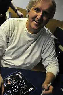 Homme habillé en blanc assis appuyé sur une table avec un stylo dans sa main gauche et une photo promotionnelle d'Avatar dans sa main droite.