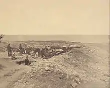 Photographie d'une position d'artillerie au sommet d'une colline rocailleuse et désolée où sont déployés deux canons derrière des talus renforcés de sacs de sable. Plusieurs dizaines d'hommes portant des tenues variées prennent la pose.