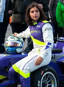 Photographie d'une femme pilote automobile blanche, brune, vue de face, en combinaison blanche, violette et jaune, assise sur une roue de monoplace, casque à la main.