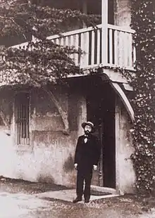 Photographie d'un homme, portant un costume et un chapeau de paille, devant une maison en partie couverte de lierre.