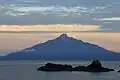 Photo couleur d'un volcan, sur une île, depuis une île voisine, sous un ciel nuageux peu lumineux.