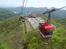 Vue aérienne couleur d'une station de téléphérique et de son parking, aménagés au sommet d'une colline boisée. Une télécabine rouge et blanche occupe une partie du premier plan, à droite. L'arrière-plan est constitué d'une chaîne de montagnes, sous un ciel bleu nuageux.