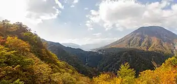 Photo couleur d'une étendue forestière aux couleurs de l'automne au premier plan, d'une montagne boisée à droite, d'un lac et d'une chute d'eau au centre. Un ciel bleu, nuageux et lumineux, en arrière-plan.