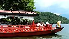 Photo couleur d'une embarcation rouge sur un lac. Quatre personnes en habit de prêtre shintō sont à bord. On distingue, en arrière-plan, un massif montagneux sous un ciel bleu nuageux.