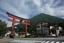 Photo couleur d'une montagne boisée sous un ciel bleu nuageux, avec, au premier plan, un portail traditionnel de sanctuaire shintō.